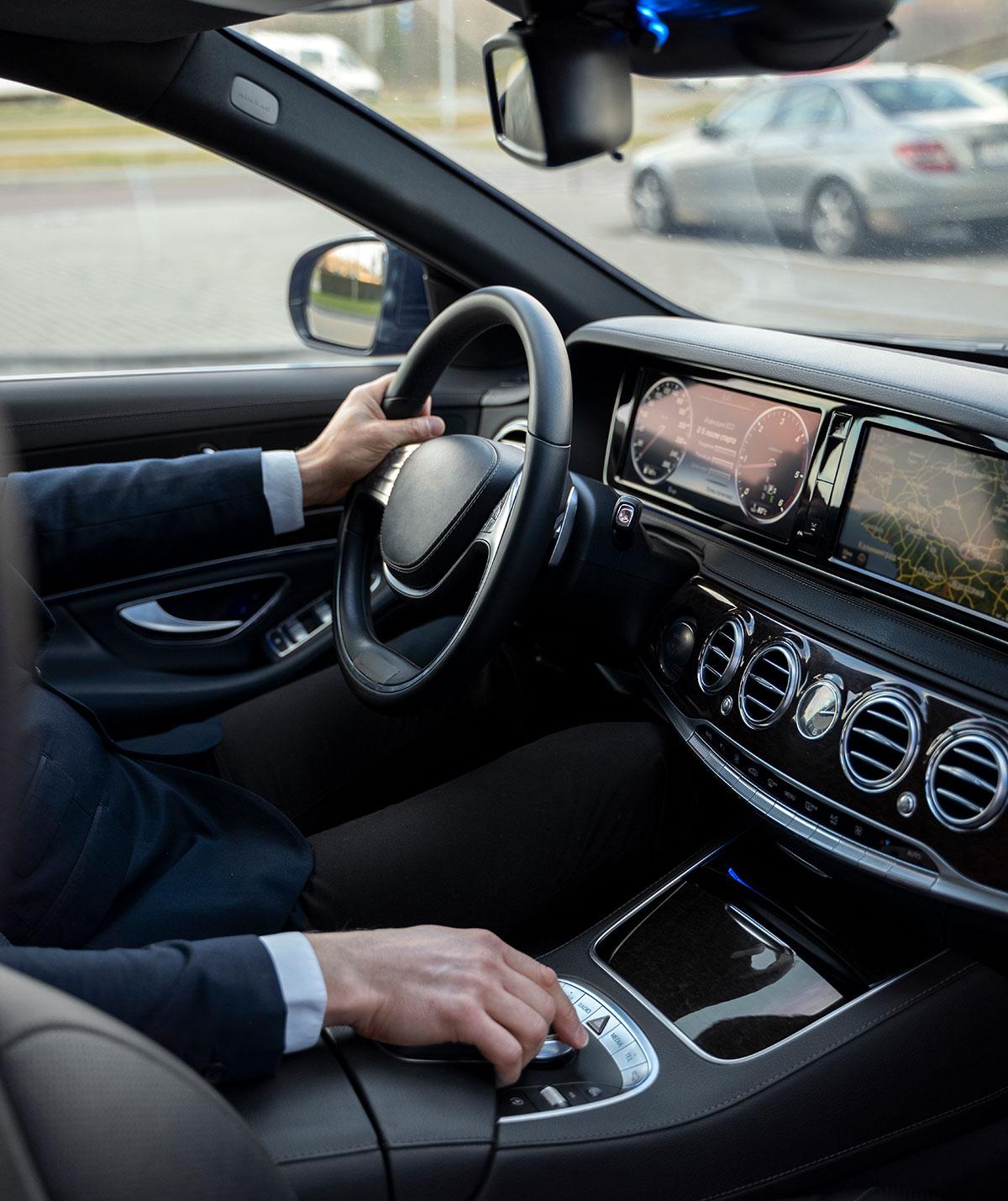 Person driving a car service vehicle, with a hand on the steering wheel and the other hand operating a control knob. The dashboard displays various information on digital screens.