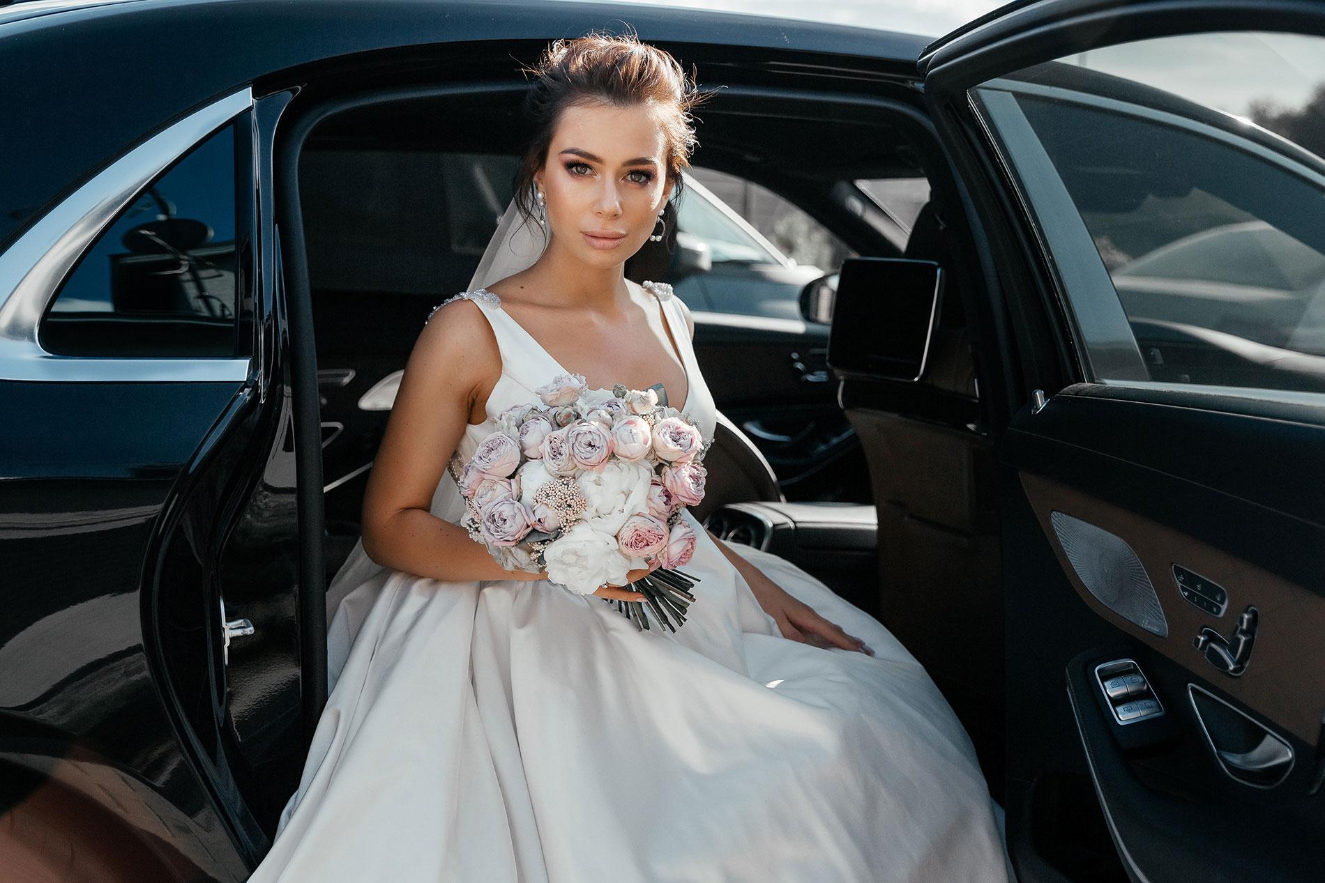 A bride in a white wedding dress holding a bouquet of flowers sits in a car service vehicle with an open door.