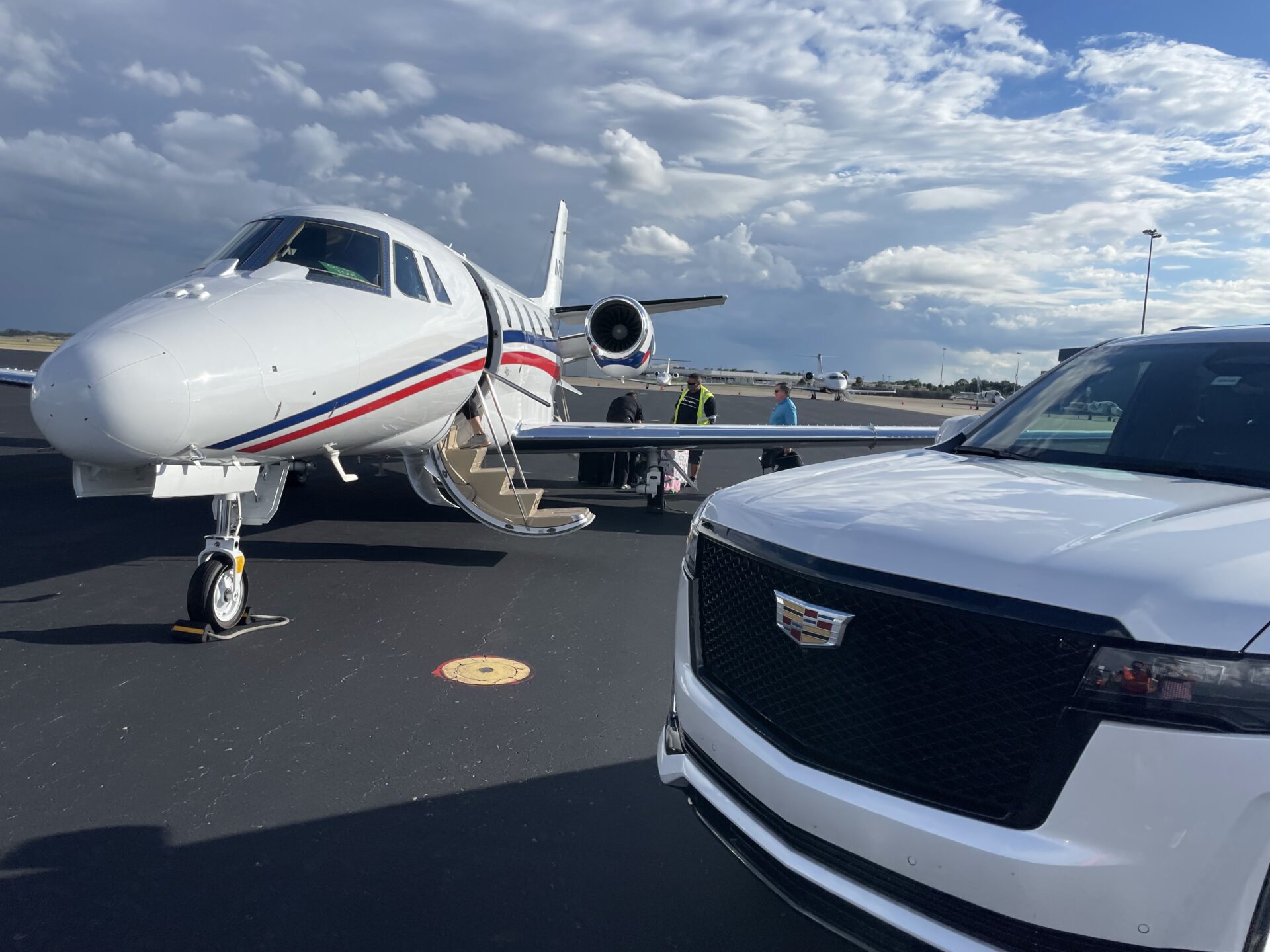 A white private jet with its stairs down is parked on an airport tarmac. A white Cadillac SUV from a car service is positioned in the foreground near the jet. Several people are near the jet.
