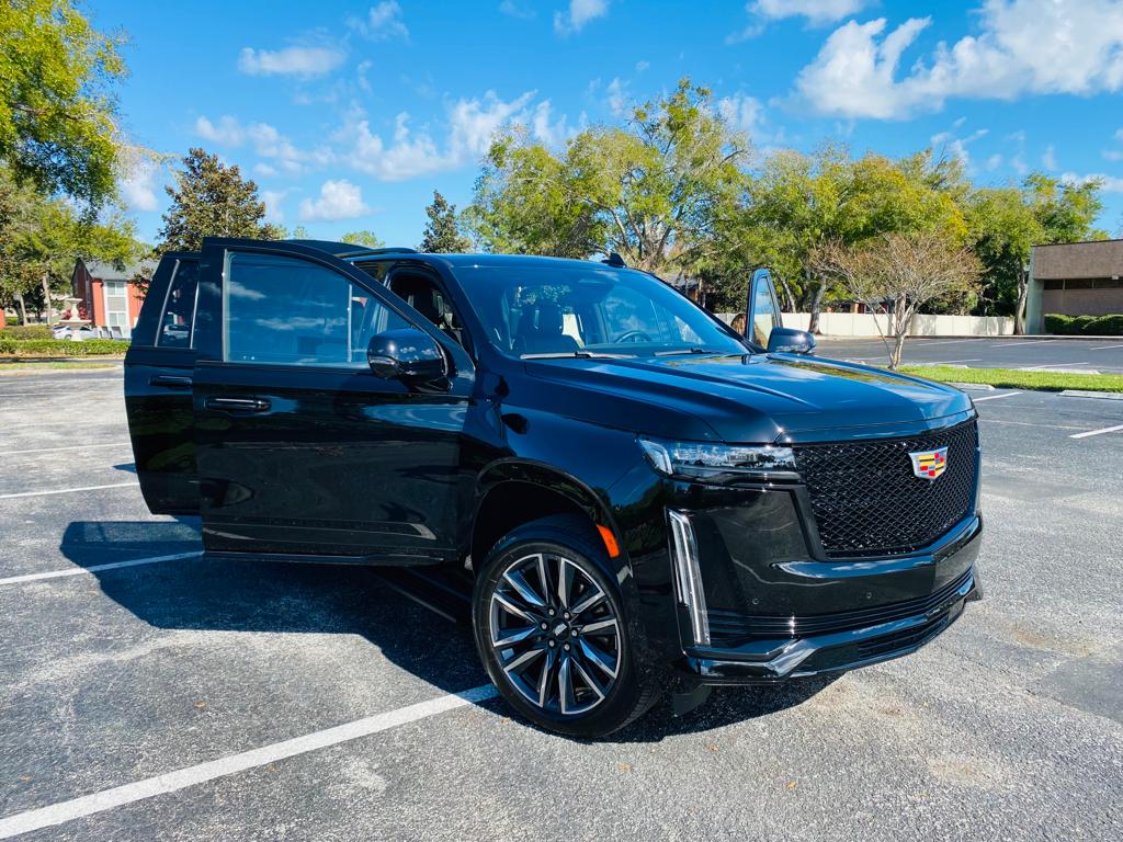 A black Cadillac SUV is parked in an outdoor lot with its driver's door open. Trees and buildings are visible in the background under a partly cloudy sky, suggesting a professional car service or Orlando limo transfer.