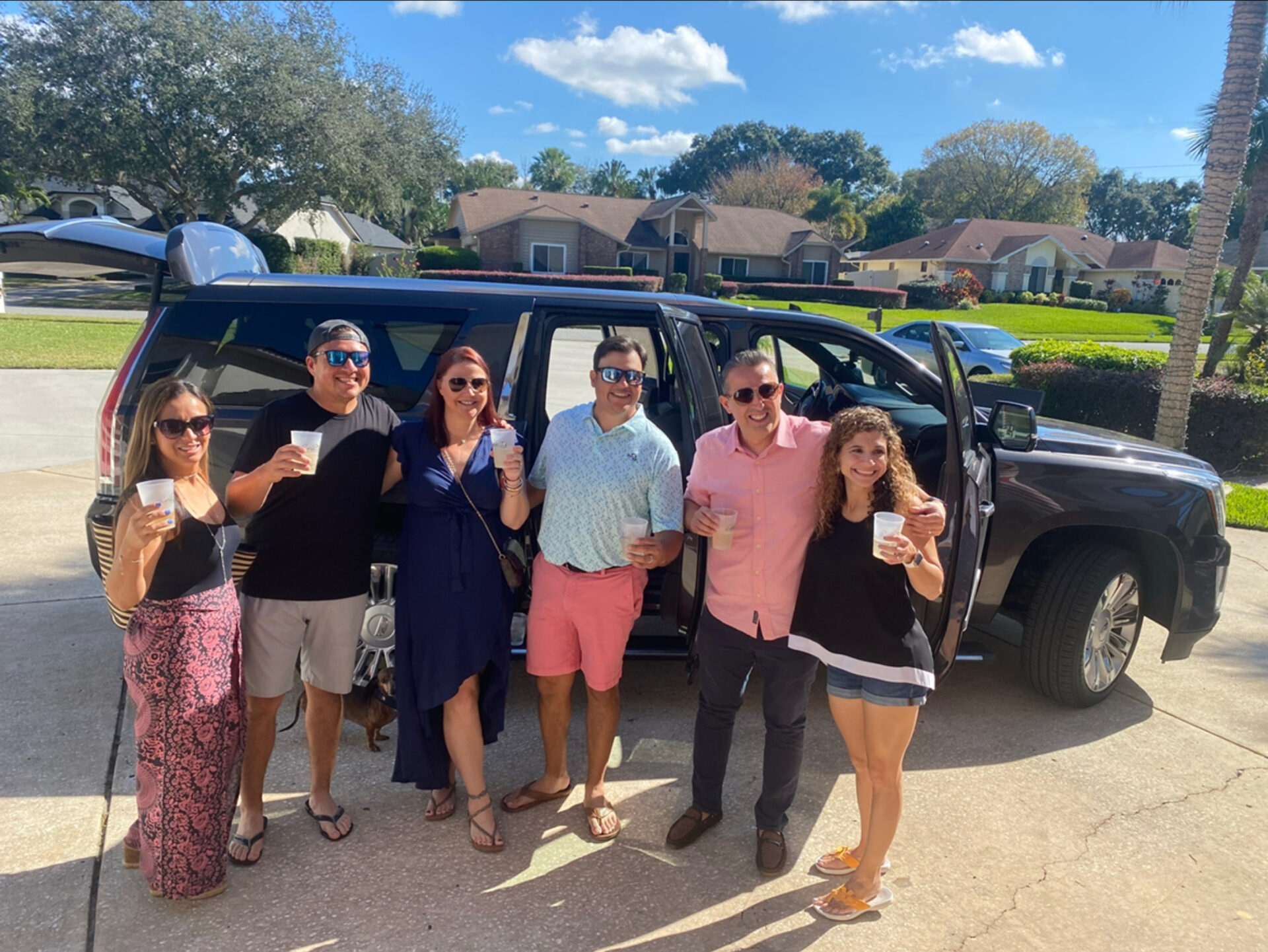 Six people stand in front of an open SUV, smiling and holding drinks, with residential houses and trees in the background on a sunny day, as if awaiting a limo transfer to their next destination.