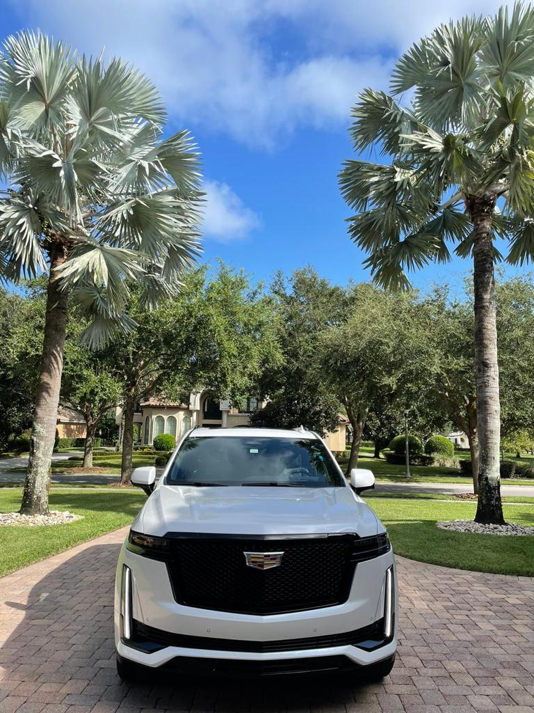 A white SUV, ideal for a comfortable airport transfer, is parked on a brick driveway under tall palm trees, with a residential house and a blue sky in the background.