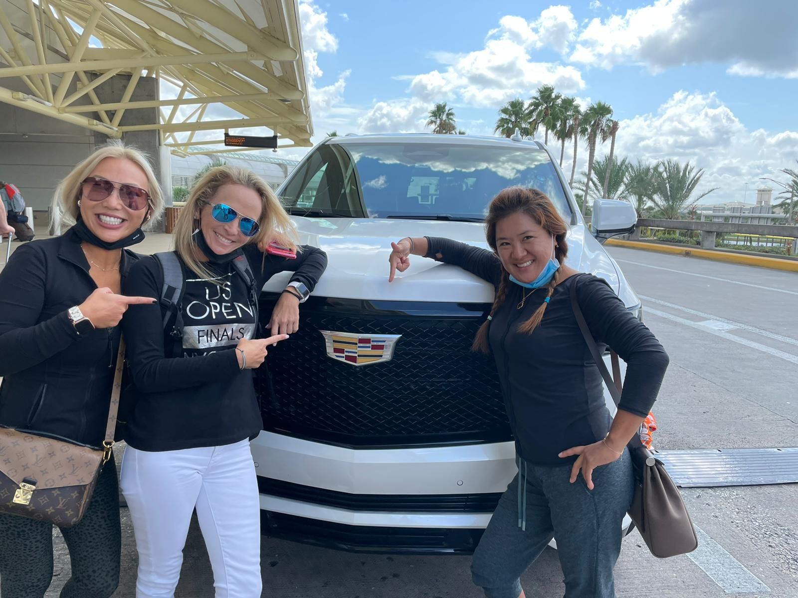 Three women smiling and pointing at a white Cadillac SUV parked under an awning, with palm trees and a cloudy sky in the background, ready for their limo transfer.