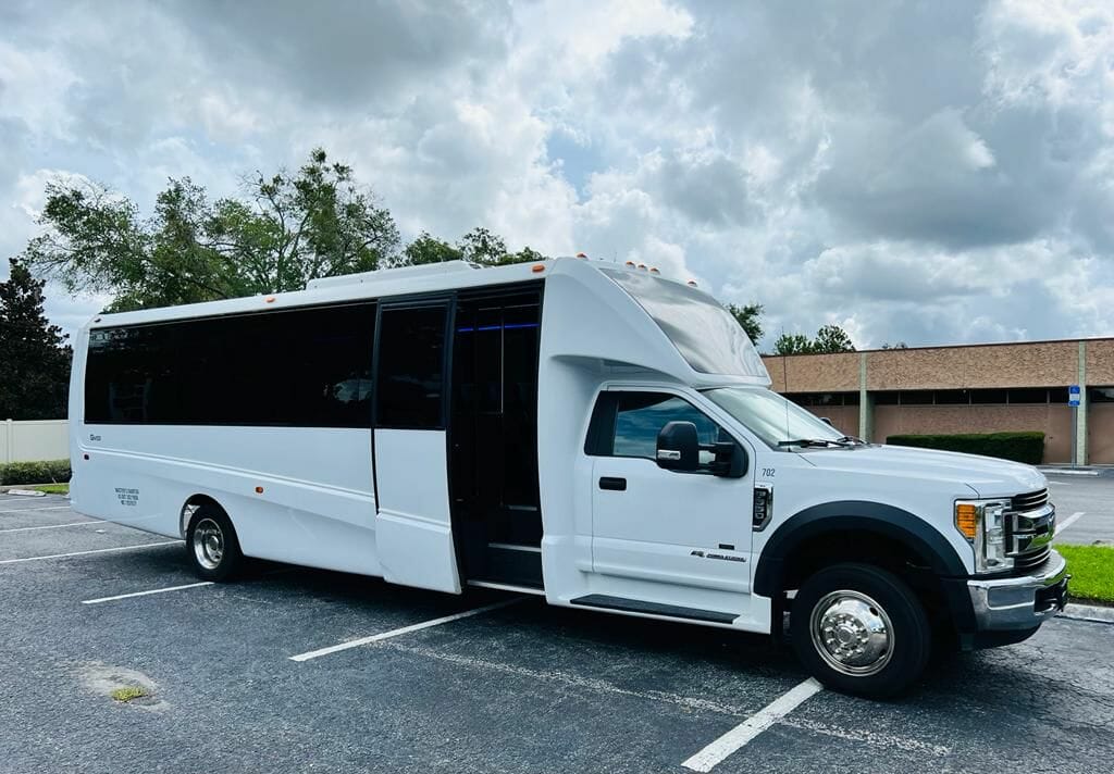 A white passenger shuttle bus associated with a car service is parked in a parking lot during the daytime, with trees and a building in the background. The bus has its door open.