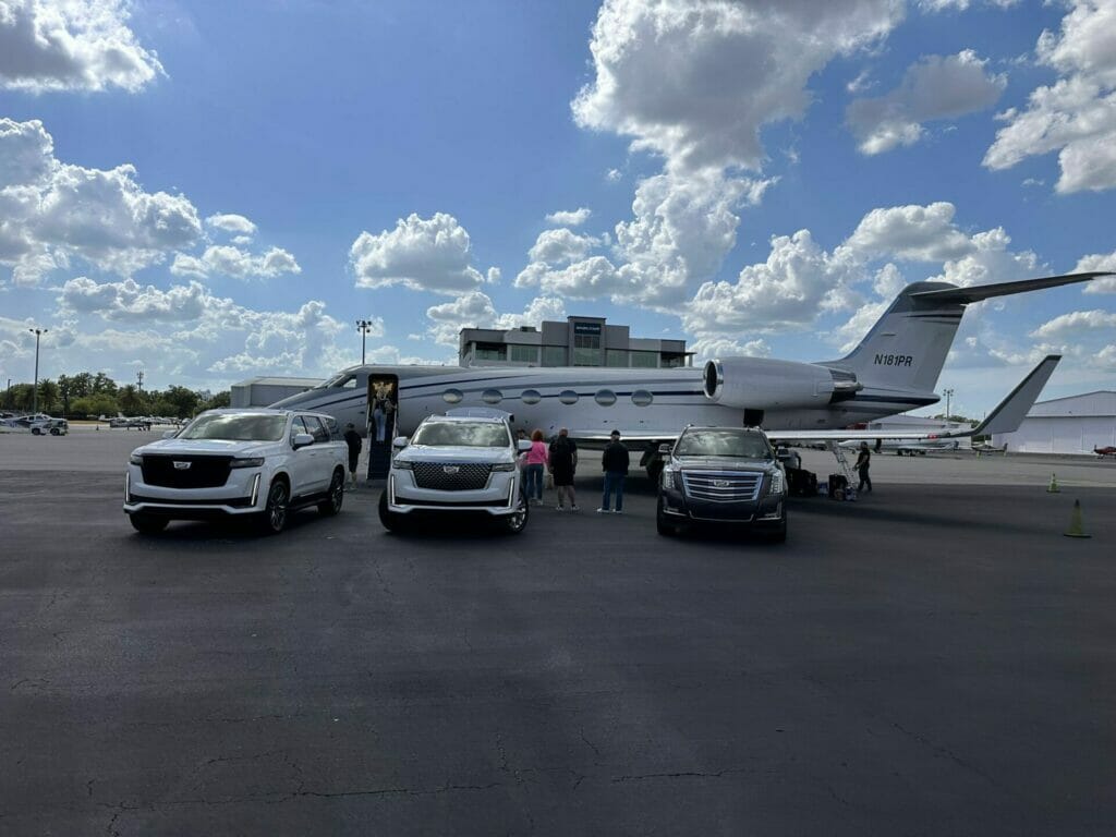 Three luxury SUVs are parked in front of a private jet on an airport tarmac, showcasing a premium limo service. Several people stand near the jet’s stairs under a partly cloudy sky.